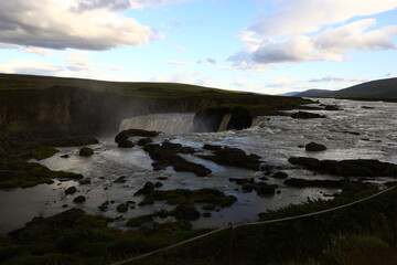 A scenic view of the upper platform of the famous Goðafoss waterfall in Iceland. The image shows flowing water, rocky edges, and the surrounding rugged landscape, captured without people.