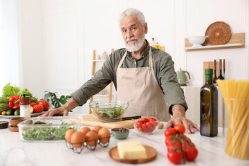 Elderly man cooking salad at white marble table in kitchen