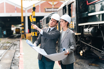 Engineers wearing safety helmets inspecting railway blueprint beside a vintage locomotive, symbolizing teamwork, innovation, and development in modern rail transportation and infrastructure projects