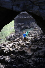 A boy in a blue shirt explores a lava cave in Iceland, holding a green jacket while standing on rugged volcanic rocks. The dark cave arch contrasts with the bright natural light outside.
