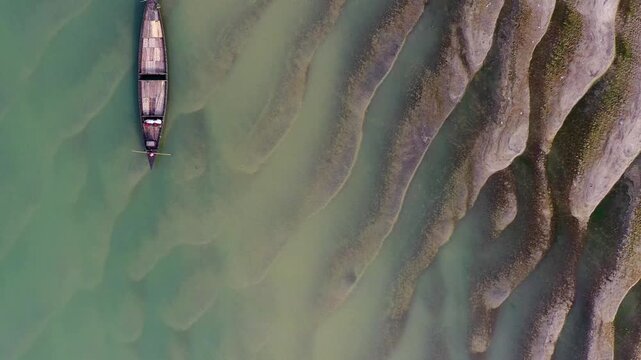 Bogura, Bangladesh - 22 August 2025: Aerial view of a slender boat cutting through the aquamarine river, contrasting with the textured, rippled shoreline.