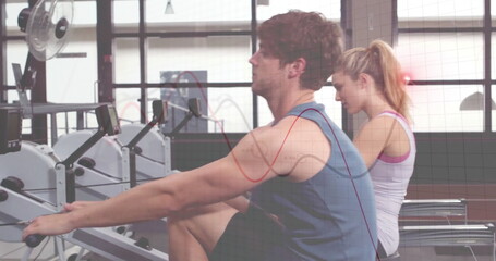 Rowing partners pushing handle bars with foot rests on rowers at fitness center, digital consoles