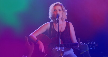 Singing teenage female musician playing acoustic guitar on club stage with mic stand and haze