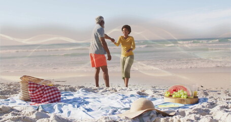 Spinning couple wearing swimwear holding hands on sandy beach with light blue picnic blanket