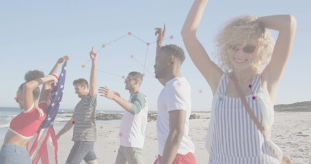 Walking five friends draping American flag and raising arms on sandy beach with rocky outcrops