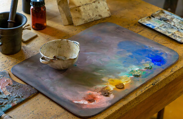 Painter’s palette with colorful oil paints, artist’s workshop table, old vessels and painting...