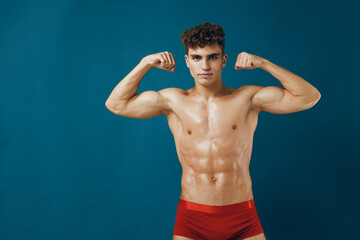 Confident young athletic man flexing muscles in red underwear against a solid blue background. Fitness and body positivity concept for health and strength.
