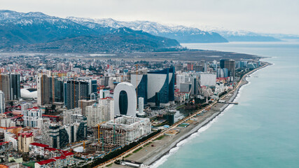 Obraz premium Aerial panorama revealing batumi cityscape, featuring contemporary skyscrapers, luxurious hotels, residential complexes lining black sea coastline, snow-dusted mountain peaks framing urban landscape