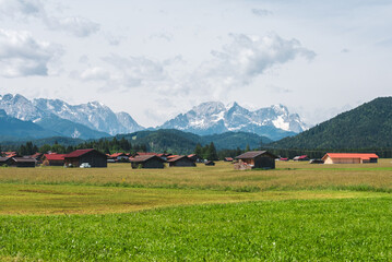 Traditional Bavarian barns and red-roofed farm buildings in green fields, with the snowy Alps rising in the background. A peaceful rural landscape in southern Germany.
