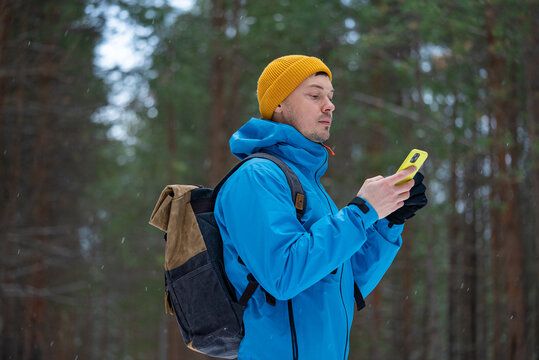 Man hiker wearing blue jacket, yellow beanie and gloves, and carrying backpack, is using smartphone while walking through snowy winter forest