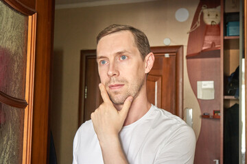 Caucasian young adult male deep in thought wearing white t shirt standing in a cozy home...