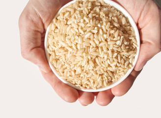 Hands holding bowl of uncooked brown rice against white background for healthy eating concept.