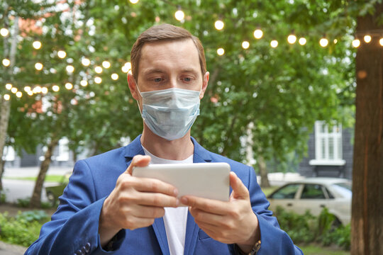 Caucasian adult male in blue suit using smartphone outdoors while wearing protective face mask.