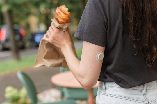 Close-up side view of a woman wearing a continuous glucose monitor standing outdoors eating a croissant in the summer
