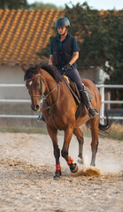 Concentrated and disciplined young female rider training trotting on her brown horse outdoors on a sandy track with a lot of dust generated by the horse's legs.