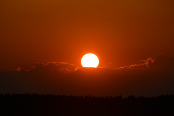 The setting sun in summer. The sun circle and the forest on the horizon.  The sun goes behind a cloud in the evening.