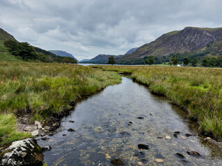 A tranquil stream winding through grassy meadows leading to a mountain lake beneath a cloudy sky.