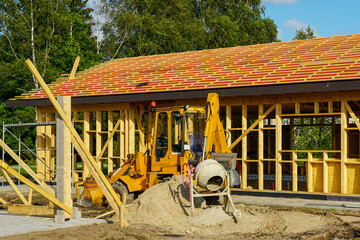 Excavator and cement mixer at wooden frame house construction site with roof beams