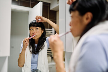 Portrait of a young asian woman cleaning brushing her teeth and using a mobile phone in front of mirror in bathroom. Dental hygiene, health and beauty concepts