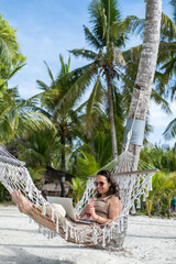 A young woman relaxes in a hammock on a beautiful beach, typing on her laptop while sipping...