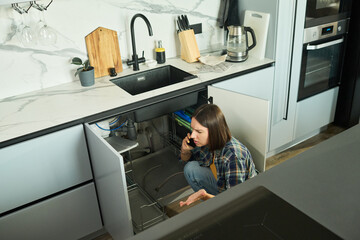 Caucasian young adult woman crouching under kitchen sink talking on smartphone appearing concerned while inspecting plumbing pipes in modern kitchen interior