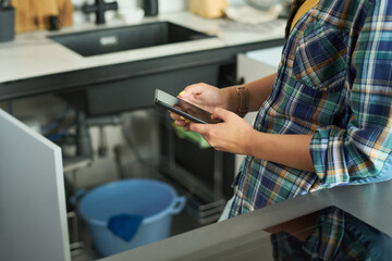 Young adult Caucasian woman standing in kitchen holding smartphone with both hands, looking at screen, wearing casual clothing, blue bucket visible under sink, partial body view