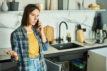 Young adult Caucasian woman standing in modern kitchen talking on smartphone with serious expression, gesturing with hand while communicating, kitchen appliances visible in background