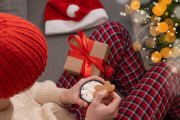 Young man in red plaid pajamas holding mug of hot cocoa topped with marshmallows and gingerbread...