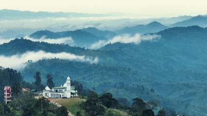 Sajek, Bangladesh - 21 August 2025: Aerial view of a striking white mosque amid green mountains, partially veiled in ethereal mist, creating a serene landscape.