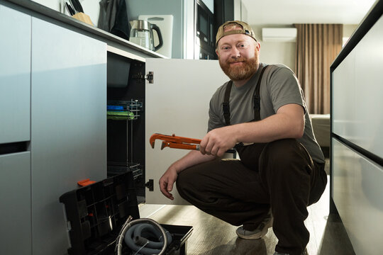 Caucasian middle aged man kneeling in modern kitchen holding pipe wrench and smiling at camera, performing plumbing repair work with open toolbox and cabinet door