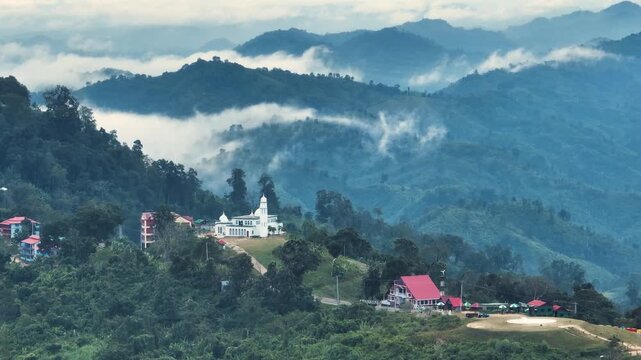 Sajek, Bangladesh - 21 August 2025: Aerial view of rolling green hills embraced by soft, low-lying clouds, with a white mosque standing prominently among the lush landscape.