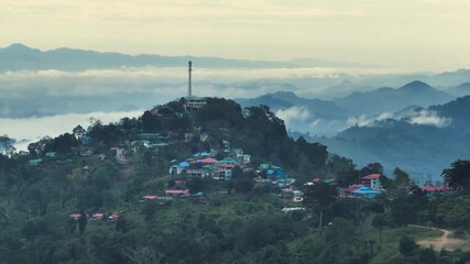 Sajek, Bangladesh - 21 August 2025: Aerial view of a village nestled on a hilltop, amidst rolling mountains and a blanket of low-lying clouds, under a hazy sky.