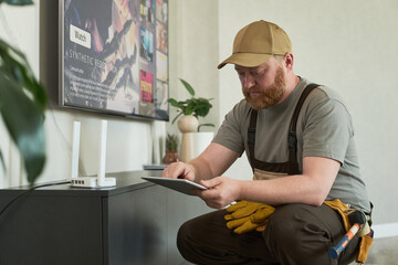 Middle aged Caucasian man with beard wearing work uniform sitting near television configuring wireless router using digital tablet, tool belt and gloves visible on side