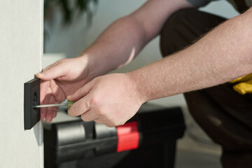 Caucasian middle aged man using screwdriver to install electrical outlet on wall, hands visible working with tool, toolbox in background, performing home maintenance task