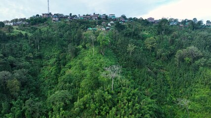 Sajek, Bangladesh - 21 August 2025: Aerial view of buildings nestled on a lush green hill, with dense trees contrasting with the clear sky above.