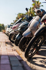 Motor scooters are parked neatly along a sunlit sidewalk. The scene captures a lively urban...