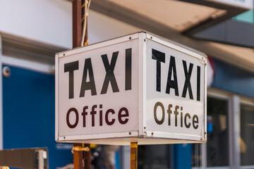 Obraz premium A taxi office sign stands out with bold letters in a vibrant urban environment. People are likely nearby, engaged in daily activities and commuting during peak hours