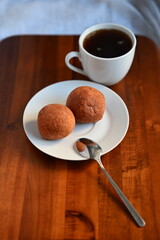 White cup of black coffee with two fried buns served on a white plate with spoon, placed on wooden table. Homemade breakfast concept.