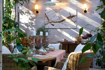 Elegant autumn table setting on a terrace with wicker chairs, candles, silver pumpkins and seasonal floral arrangements. Cozy rustic atmosphere with natural light and greenery