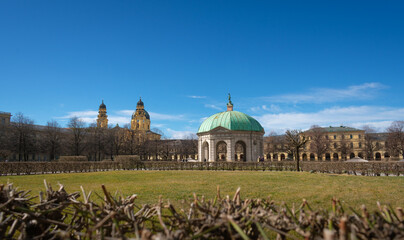 The Diana Temple (Dianatempel) in the Hofgarten, Munich, Germany, with the baroque towers of the Theatine Church in the background. A scenic blend of garden, culture, and historic architecture.