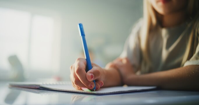 Close Up Shot of Young Girl Sitting at the Desk, Writing Exam, Doing Task or Drawing in Notebook. In the Background, Blurred Children During School Test in Bright Classroom of Modern Primary School.