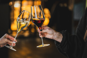 Two hands clinking wine glasses with red and white wine in warm ambient light. Cozy celebration moment in a bar or restaurant, symbolizing friendship, toast, and enjoyment