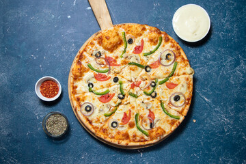 Delicious Veggie Feast Pizza with Mushrooms, Olives, Onion, Tomato and Bell Peppers isolated on wooden cutting board on a Dark Blue Background top view of fast food