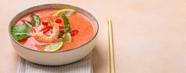 Creamy flavourful tom yum soup, a popular dish of Thailand, is served in a bowl, garnished with fresh shrimp, lime wedges, herbs, and chili peppers, accompanied by chopsticks on a linen napkin, copy
