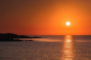 A vibrant orange sunset over a calm ocean, the sun near the horizon, casting a warm glow on the water and silhouetting a rocky shoreline