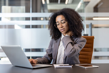A tired African American young businesswoman sits at an office desk, rubs her neck with her hand,...