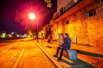 Lonely man sitting on a bench next to a streetlight on an empty street at night, in Parral,...