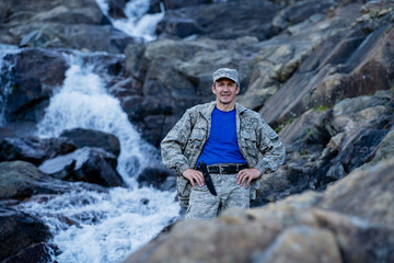 A hunter in camouflage stands near a waterfall among rocks