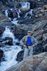 A contented Hunter in camouflage with a knife stands near a waterfall among stones
