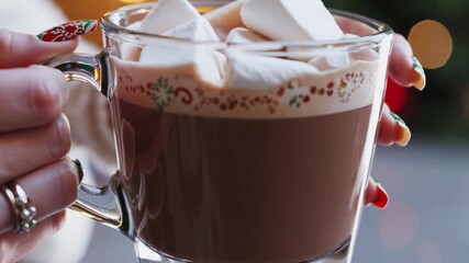 Female hands exhibiting bright festive nail art design cradling glass mug filled with hot chocolate beverage and marshmallows during winter holiday season - Powered by Adobe